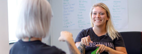 To kvinder taler tegnsprog i et undervisningslokale. Den ene har front mod kameraet og smiler.