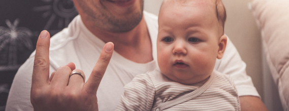 En baby sidder på skødet af sin far og kigger på hans hånd, der laver et tegn