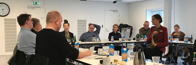 Deltagere ved møde sidder ved borde i hestesko med en tegnsprogstolk i midten.