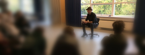 En mand i sort tøj og med hat på sidder på en stol foran en gruppe unge. De er sløret på billedet.