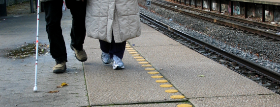 Benene af to personer, den ene med hvid stok, som går på en togperron
