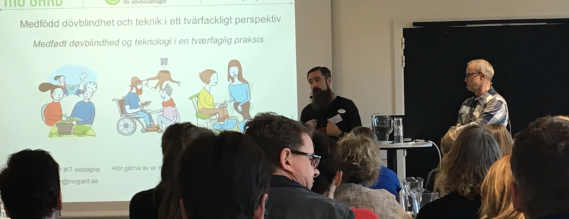 To mænd står og forelæser ved et lærred med en PowerPoint på.