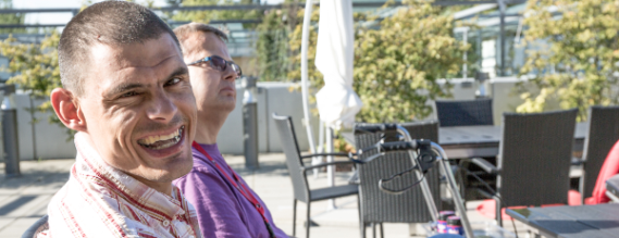 En ung spastisk mand griner ind i kameraet. Han sidder sammen med en anden mand på en terrasse med grønne træer i baggrunden.