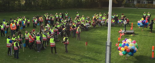 En stor gruppe beboere står iført gule veste i CFD's aktivitetspark, hvor de er i gang med opvarmning. Der er også balloner.