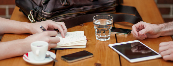 To personer sider overfor hinanden ved et bord. Man kan kun se deres hænder - den ene person skriver i en lille bog. På bordet er der en kaffekop, et glas, to iPhones, en mini iPad og en taske.